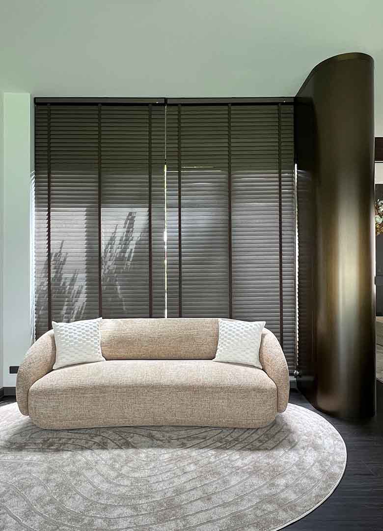 Modern sitting nook at AIRI Km.5 by Elementi Interiors, featuring a beige curved fabric sofa on a textured oval rug, dark wooden Venetian blinds, and a smooth rounded bronze-steel architectural column enclosure.