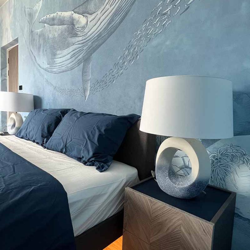 Modern coastal kids bedroom at AIRI KM.5 by Elementi Interiors, featuring an Inkiostro Bianco whale wallpaper mural, custom navy platform bed with under-bed LED lighting, herringbone wood nightstand, and ceramic table lamp.
