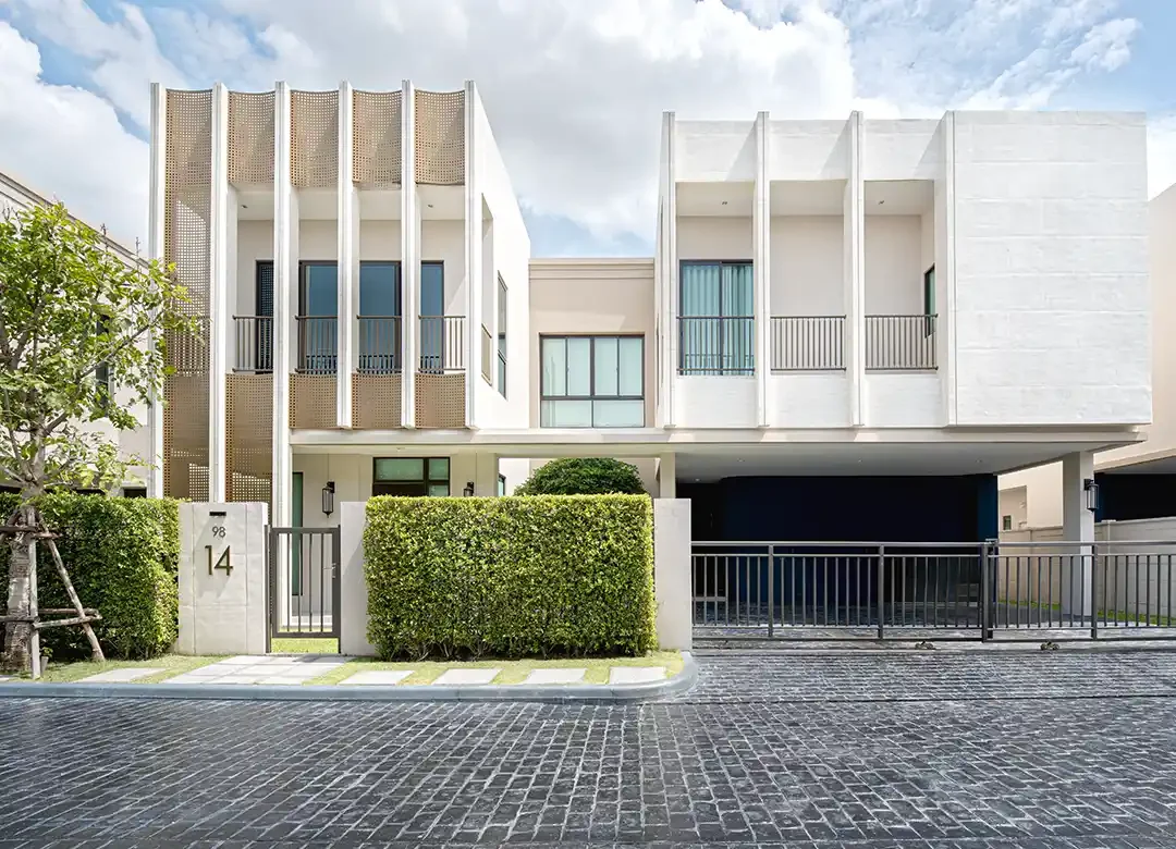 Exterior front view of a modern two-story luxury house at AIRI Km.5 featuring decorative vertical fins, woven lattice screens, landscaped hedges, and a paved driveway.