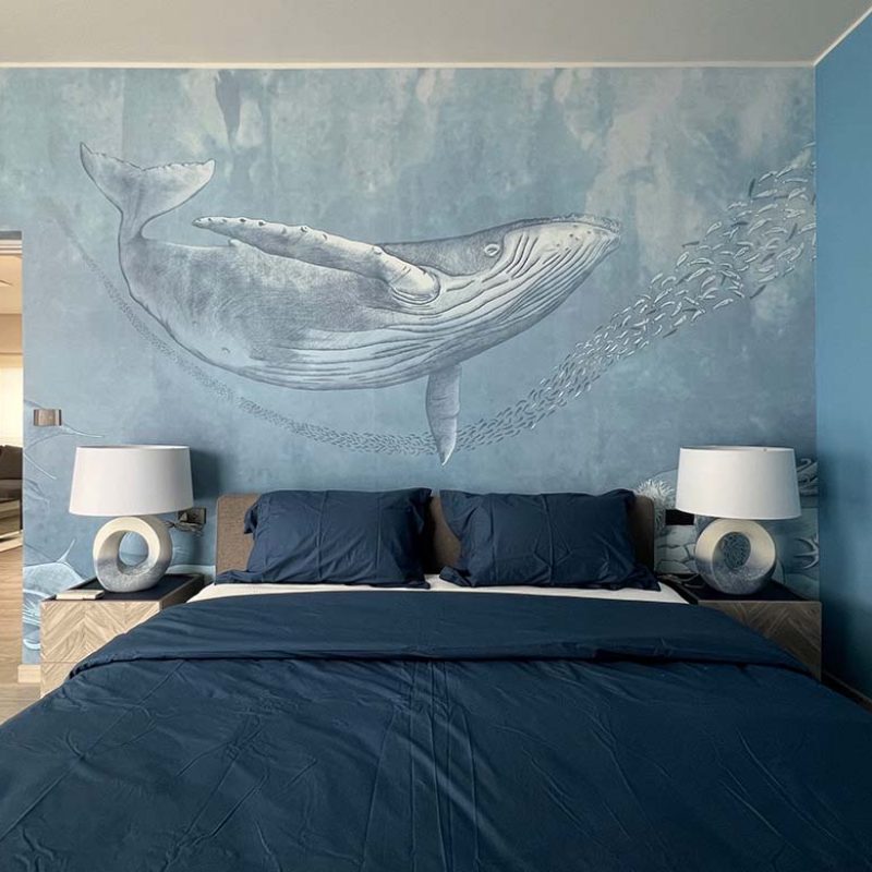 Centered view of a modern boy's bedroom in the AIRI Km.5 project featuring a blue ocean whale wallpaper mural by Inkiostro Bianco behind a bed with navy bedding and ceramic nightstand lamps.