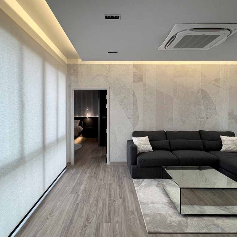Spatial view of a minimalist media playroom in the AIRI Km.5 residence highlighting a deep charcoal L-shaped couch, textured geometric accent wall, drop-down ceiling panel with perimeter LED illumination, and light-filtering window blinds.