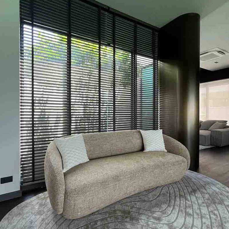 Modern sitting area in the AIRI Km.5 project by Elementi Interiors featuring a beige organic curved sofa, textured wave-patterned rug, floor-to-ceiling dark wooden blinds, and a metallic accent column.