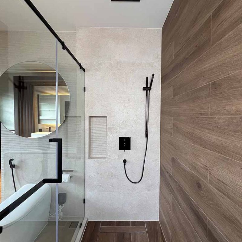 Modern glass walk-in shower room in the AIRI Km.5 project featuring wood-look porcelain floor and wall tiles, a recessed shower niche, flush ceiling rain shower head, and matte black fixtures by ROCA Spain.