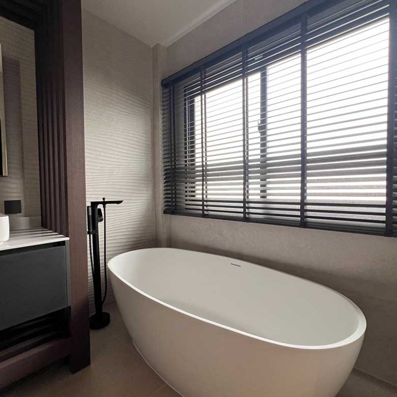 Freestanding oval bathtub in the AIRI Km.5 project featuring a floor-mounted matte black faucet, dark wooden Venetian blinds over a large window, and wave-textured ceramic wall tiles by Porcelanosa Spain.