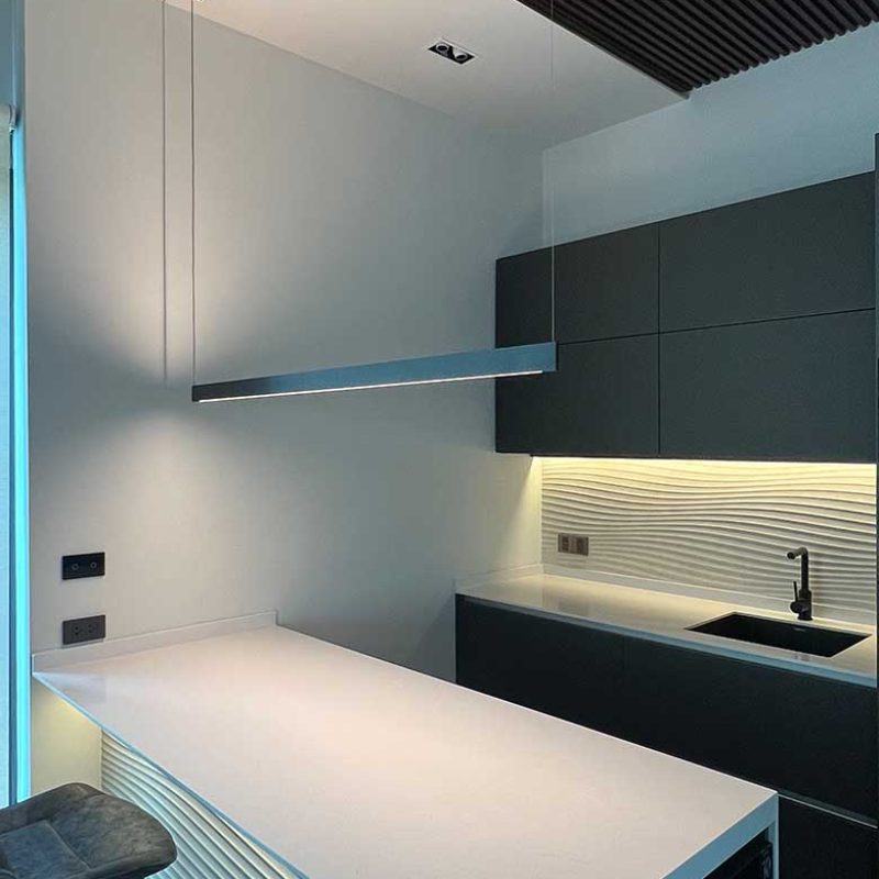 Warmly illuminated modern bespoke kitchen at AIRI Km.5 by Elementi Interiors, featuring a white Porcelanosa Samui 3D wave fascia, matte charcoal cabinetry, and the razor-thin, minimalist black linear Sottile suspension lamp by Davide Groppi suspended above.