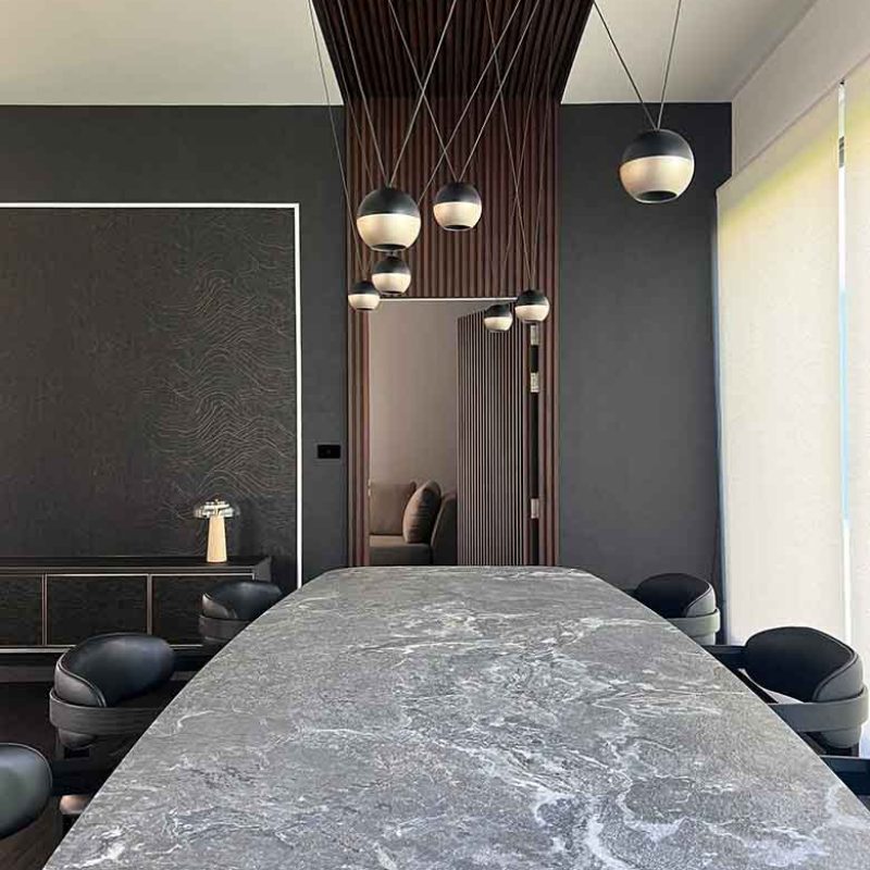 Dramatic close-up down the length of a marble-effect ceramic dining table top Tyron Keramik by Cattelan Italia, showing suspended dual-tone orb pendant lights and an open hidden slatted door within a dark wood baffle wall feature at AIRI Km.5 by Elementi Interiors.
