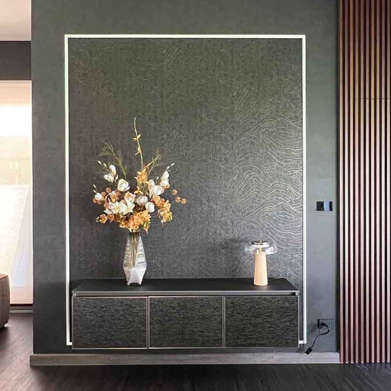 A floating dark console cabinet with bronze-framed doors upholstered in Jim Thompson fabric, set against a framed dark wall featuring a gold-patterned wallpaper, styled with a floral vase and table lamp at AIRI Km.5 by Elementi Interiors.