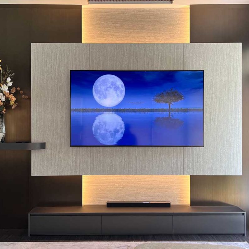 Symmetrical straight-on view of a custom TV media wall by Elementi Interiors at AIRI Km.5, featuring an accurated travertine laminate paneling with warm integrated LED backlighting, brushed bronze cladding, floating shelf, and low-profile console.