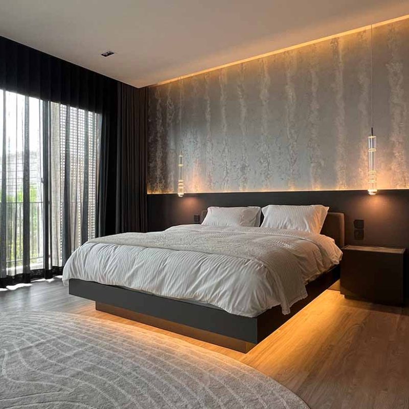 Angled view of the primary bedroom suite at AIRI Km.5 by Elementi Interiors, featuring a floating bed with warm LED perimeter lighting, custom hand-painted silver metallic accent wall with glass crystal dust, and modern hanging pendants.