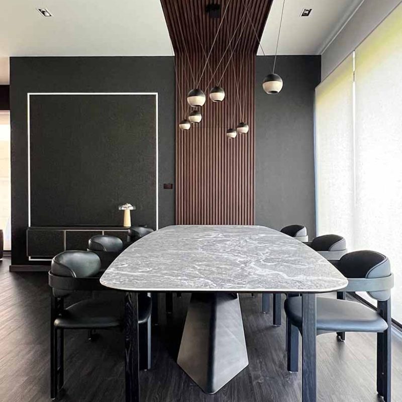 Symmetrical shot of a modern dining room at AIRI Km.5 by Elementi Interiors, featuring a Cattelan Italia Tyron Keramik dining table, dark leather chairs, spherical pendant lights, and a dark wood slat ceiling baffle.