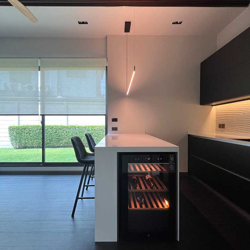 Side-profile view of a bespoke white kitchen island at AIRI Km.5 by Elementi Interiors, featuring an integrated glass-door wine cooler cellar, glowing LED Davide Groppi Sottile pendant light, dark matte cabinetry, and large sliding glass doors overlooking a landscaped garden.