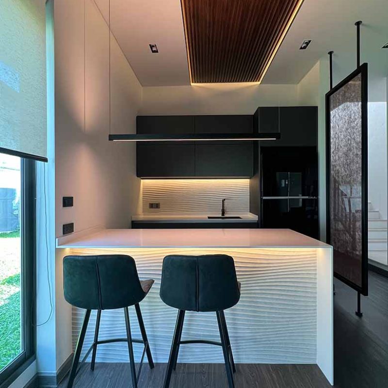 Symmetrical, eye-level shot of a modern kitchen at AIRI Km.5 by Elementi Interiors, showcasing two dark grey upholstered bar stools positioned at a counter with illuminated Porcelanosa Samui 3D wave ceramic tiles, a Davide Groppi Sottile linear suspension lamp, matte charcoal upper cabinets, and a central composite wood slat ceiling detail.