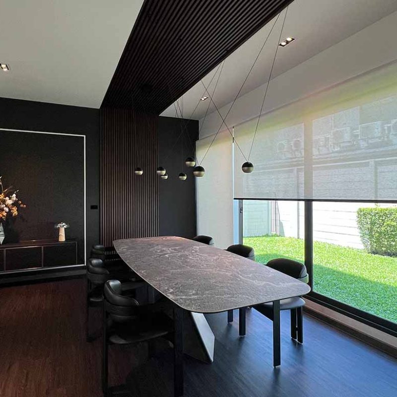 Angled view of the formal dining space at AIRI Km.5 by Elementi Interiors, featuring a Cattelan Italia Tyron Keramik table beside floor-to-ceiling glass windows overlooking a green garden, with suspended pendant lights and a floating credenza.