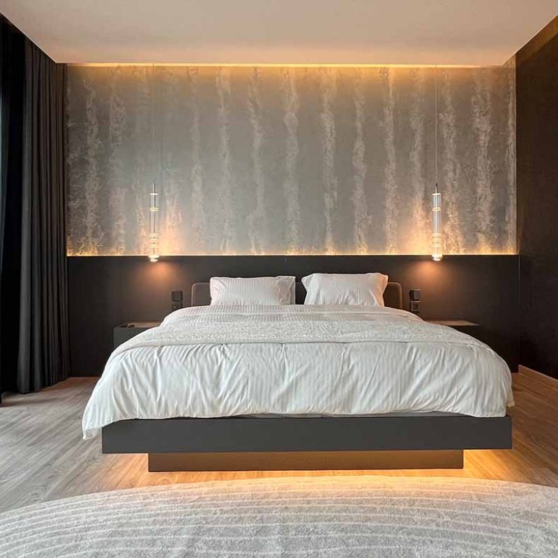 Symmetrical straight-on view of the master bedroom at AIRI Km.5 by Elementi Interiors, showcasing a floating bed with under-bed LED glow, dark headboard paneling, hanging glass pendants, and a hand-painted glass metallic feature wall.