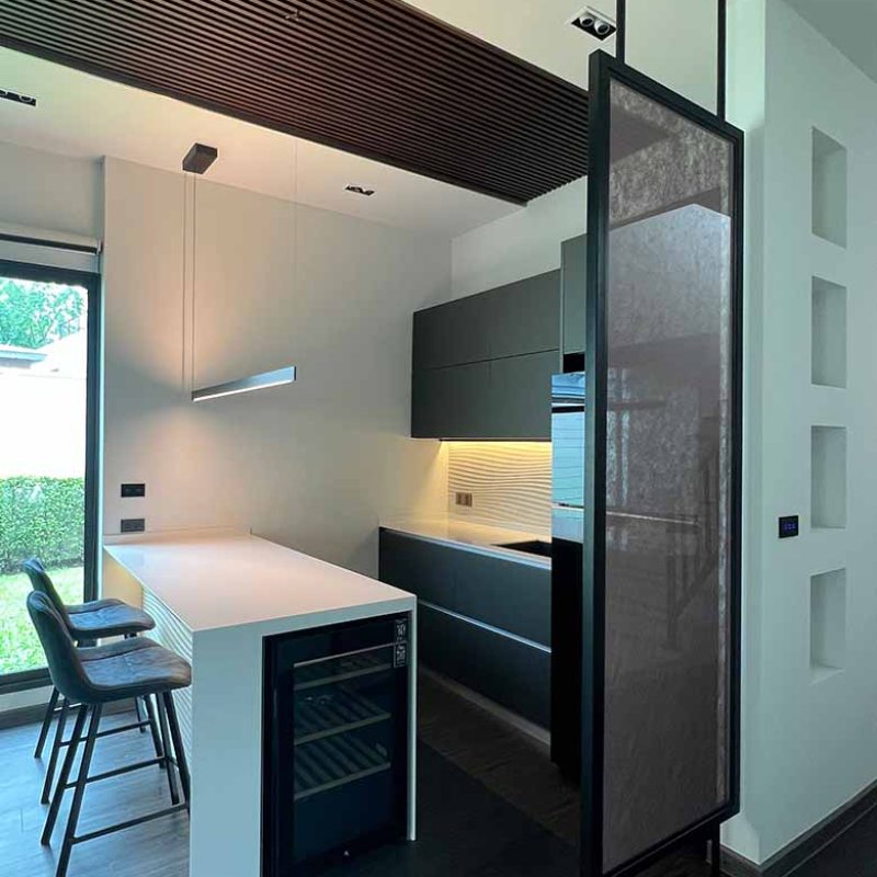 Modern interior showing a bespoke steel frame entrance partition with fabric-laminated glass, custom recessed wall niches, dark kitchen cabinetry, and a slatted wood ceiling feature.